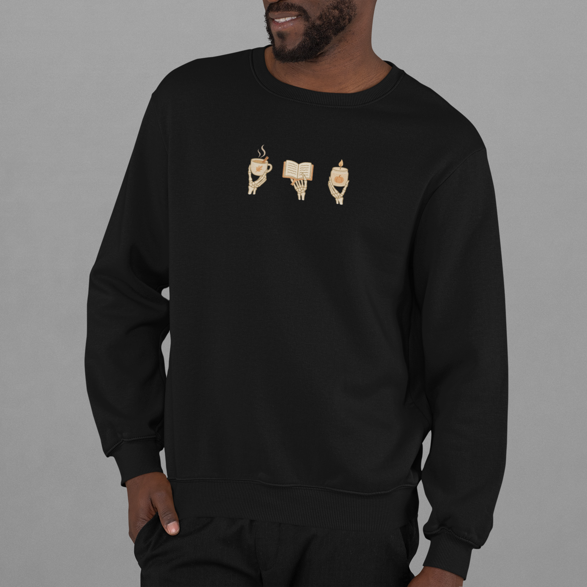 Black crewneck sweatshirt with minimalist 'Cozy Bones' graphic, featuring illustration of skeleton hands holding coffee, book, and candle. Halloween & seasonal graphic tees and hoodies.