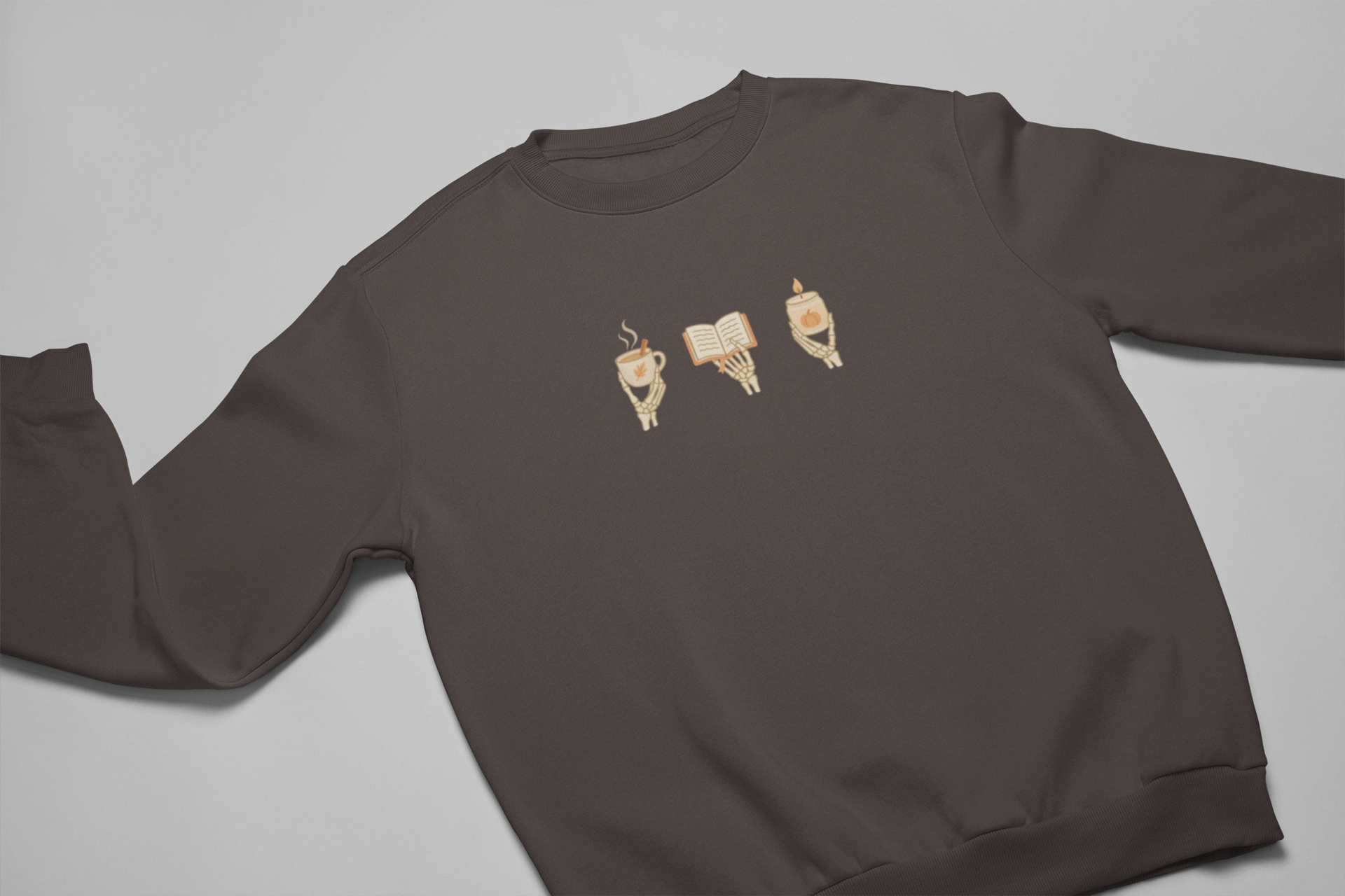 Brown crewneck sweatshirt with minimalist 'Cozy Bones' graphic, featuring illustration of skeleton hands holding coffee, book, and candle. Halloween & seasonal graphic tees and hoodies.