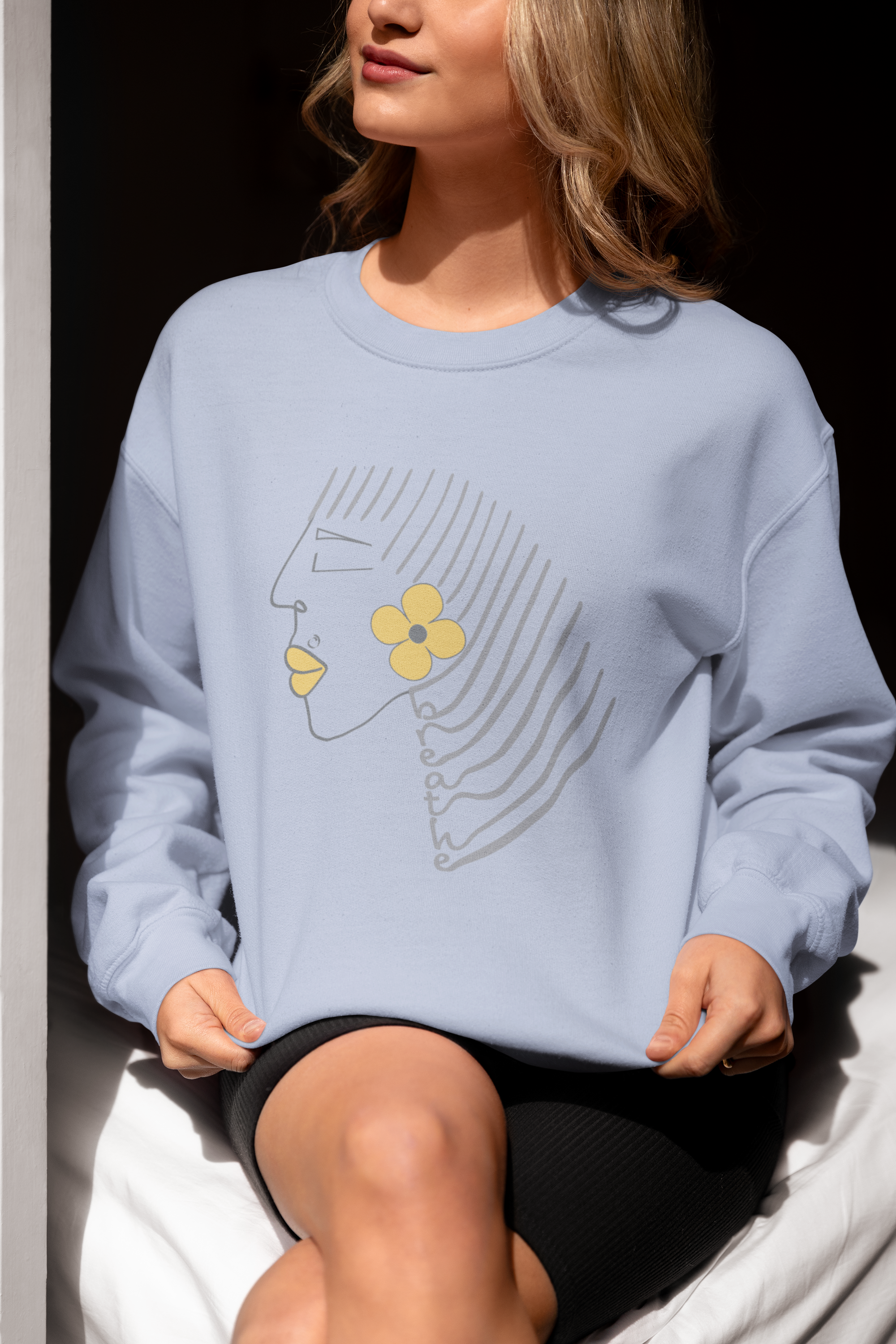 Light blue crewneck sweatshirt with minimalist Breathe graphic, featuring abstract face line art, yellow flower, and text along flowing hair lines. Positive and motivational graphic tees.