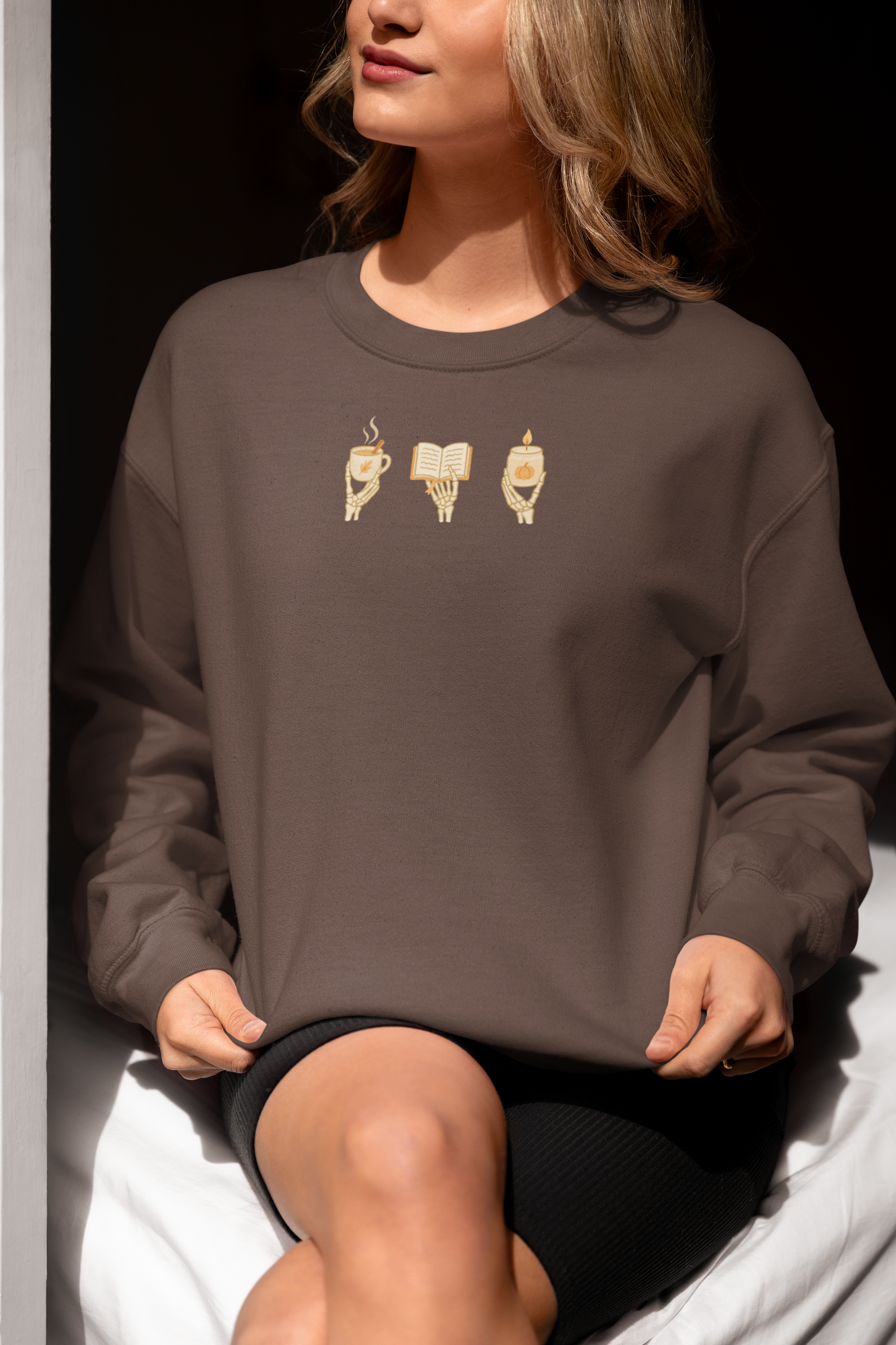 Brown crewneck sweatshirt with minimalist 'Cozy Bones' graphic, featuring illustration of skeleton hands holding coffee, book, and candle. Halloween & seasonal graphic tees and hoodies.